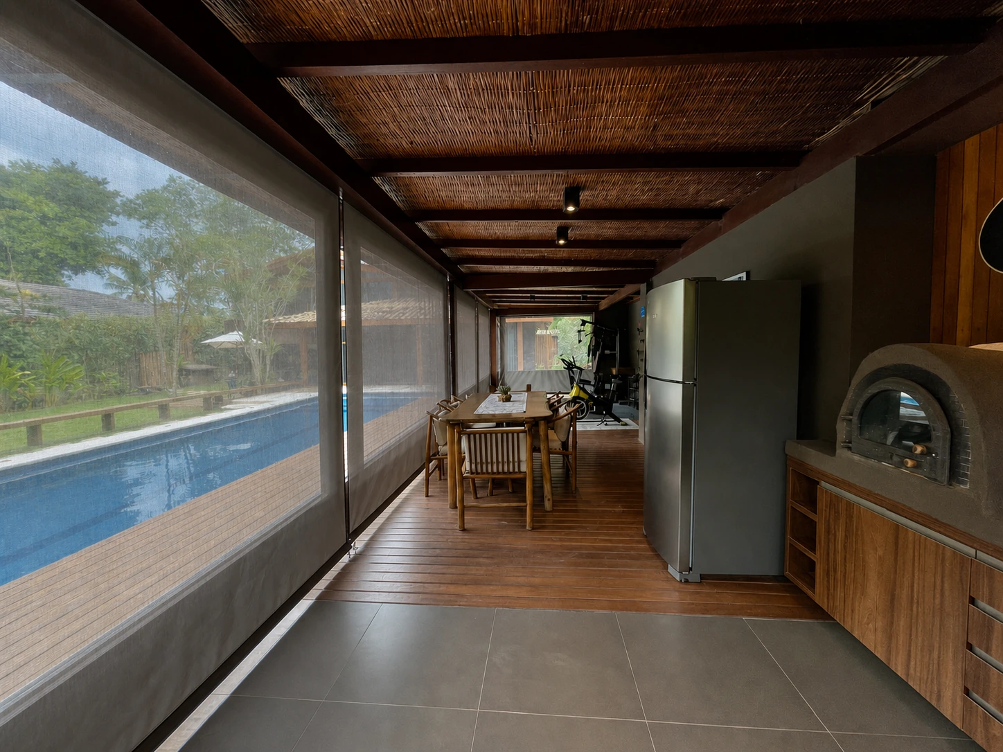Área gourmet de Casa Paraíso protegida por toldos laterales, con la piscina a la izquierda y el horno de barro a la derecha.