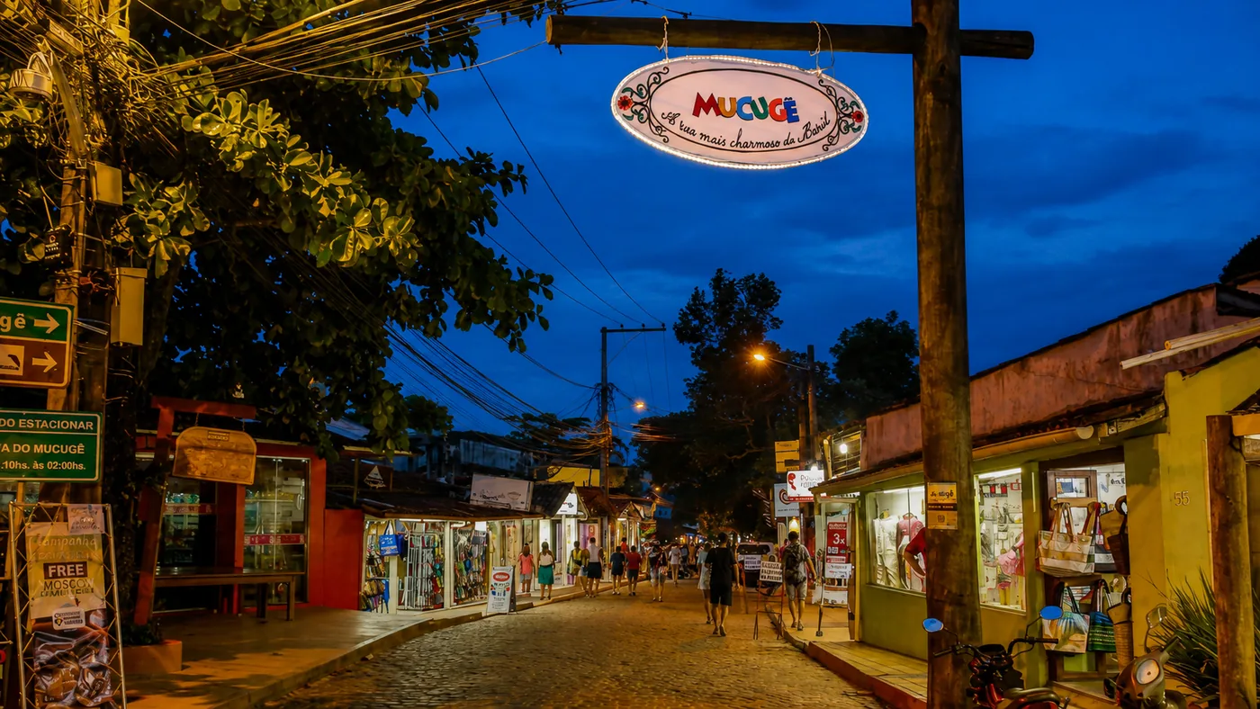 Rua do Mucugê iluminada ao anoitecer, com a placa suspensa e as lojas do vilarejo de Arraial d'Ajuda